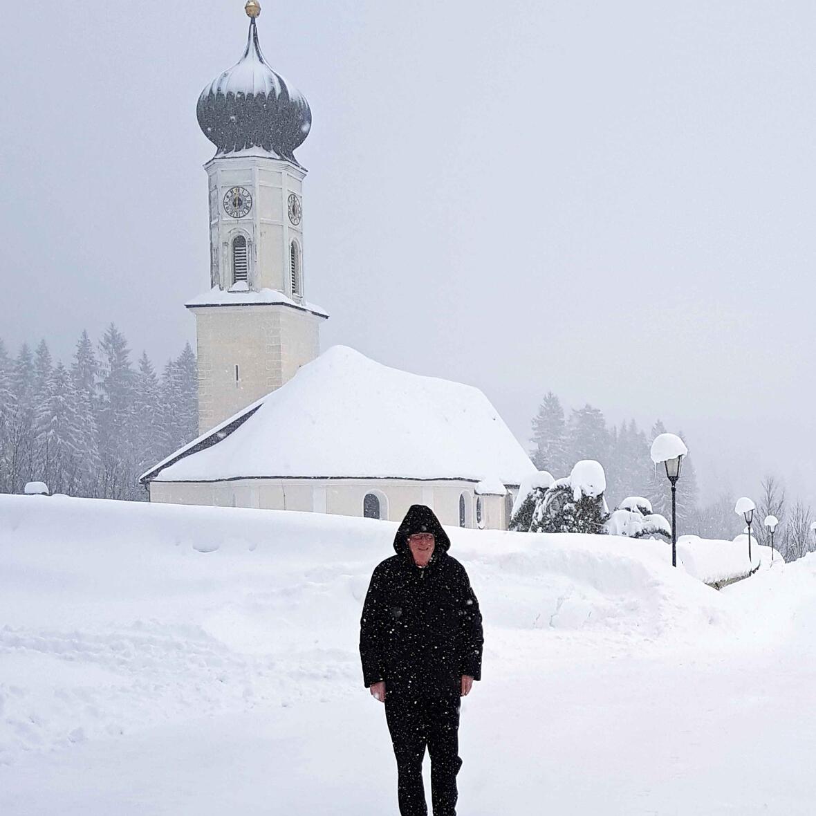 Nimmt die Situation mit Humor und einer gesunden Portion Gelassenheit: Pfarrer Willi Milz vor seiner von Schneemassen umgebenen Pfarrkirche St. Nikolaus. (Foto: privat)