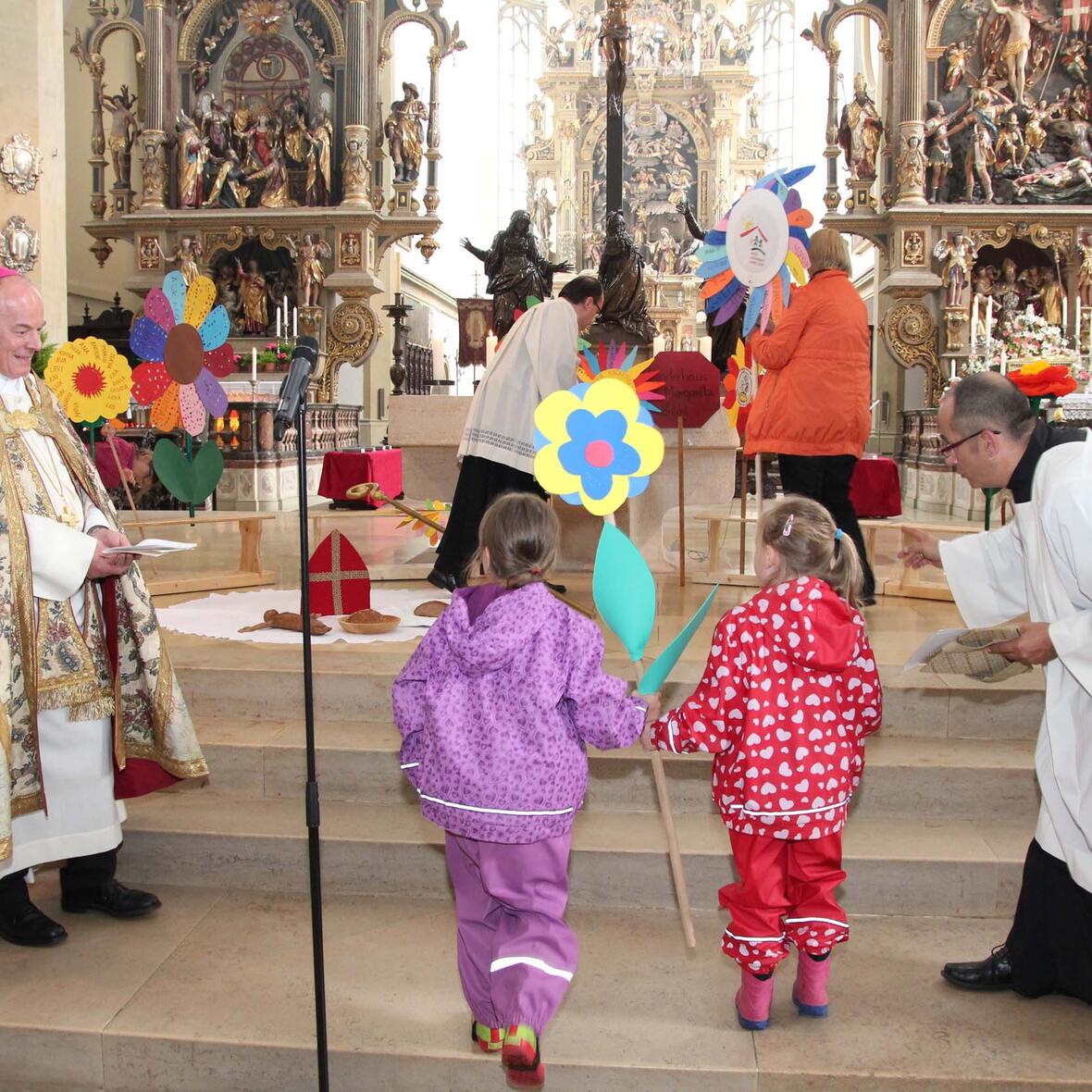 Kinder bringen ihre gebastelten Blumen als Gastgeschenk ihrer Kindertageseinrichtungen zum Altar. Weihbischof Losinger (l.) hatte ihnen erzählt, wie sehr sich der Bischof Ulrich um die armen und hungrigen Menschen gesorgt hatte. (Foto: Caritas)