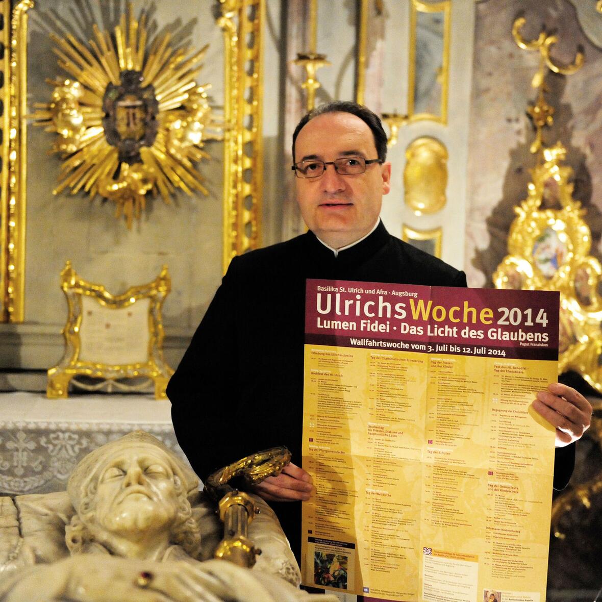 Stadtpfarrer Lothar Hartmann mit dem Plakat zur Ulrichswoche am Grab des hl. Ulrich (Bild: pba/Nicolas Schnall).