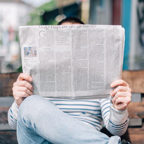 Ein Erwachsener sitzt auf einer Holzbank und liest in einer aufgeklappten Zeitung