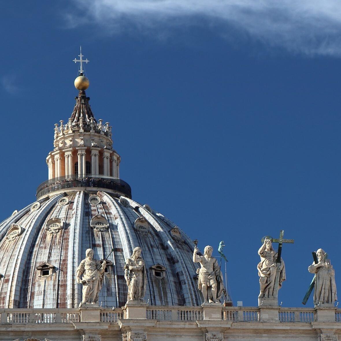 Kuppel des Petersdoms mit blauem Himmel und einigen steinernen Apostelfiguren auf den Kolonaden