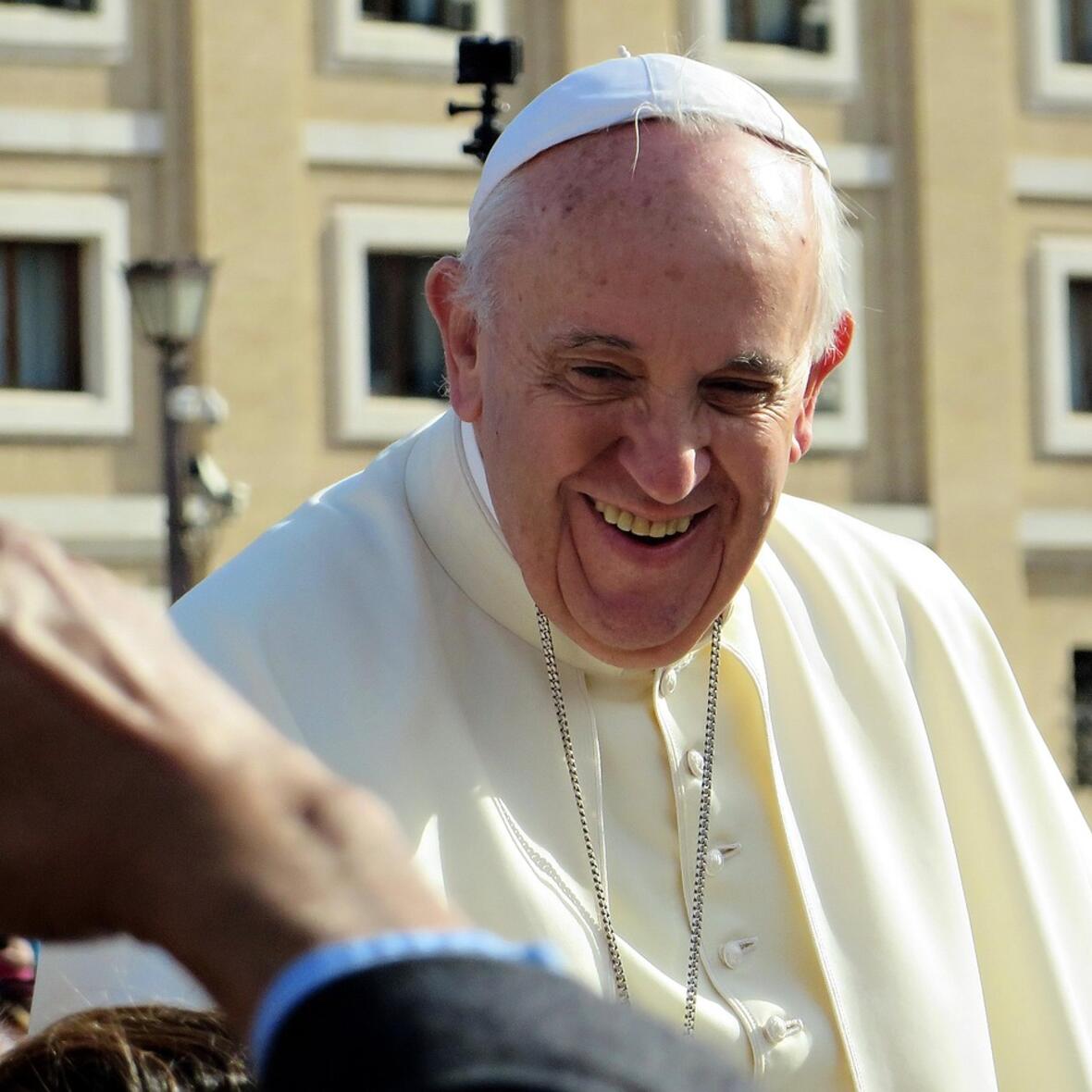 Papst Franziskus lacht bei einer Begegnung mit Menschen. Am Rand ist ein Arm zu sehen, der ein Foto vom Papst machen möchte.