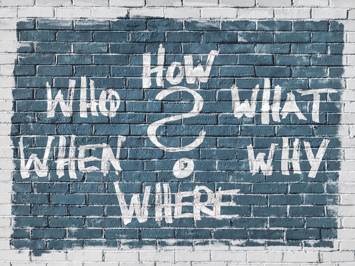 Auf einer Mauer befindet sich ein blaues Rechteck. In diesem ist im Zentrum ein weißes Fragezeichen. Außenherum finden sich in weißer Schrift die englischen Frageworte: Why, who, when, where, why, what