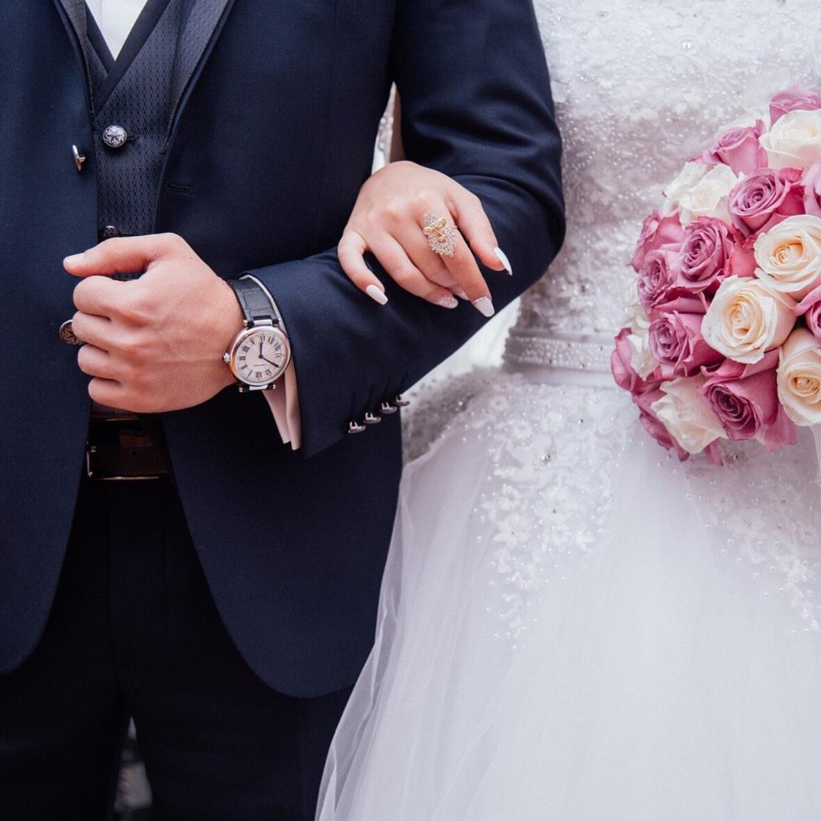 Ein Mann in einem blauen Anzug der eine Frau im Brautkleid untergehakt hat. Sie hält einen Brautstrauß mit Rosen in der linken Hand. 