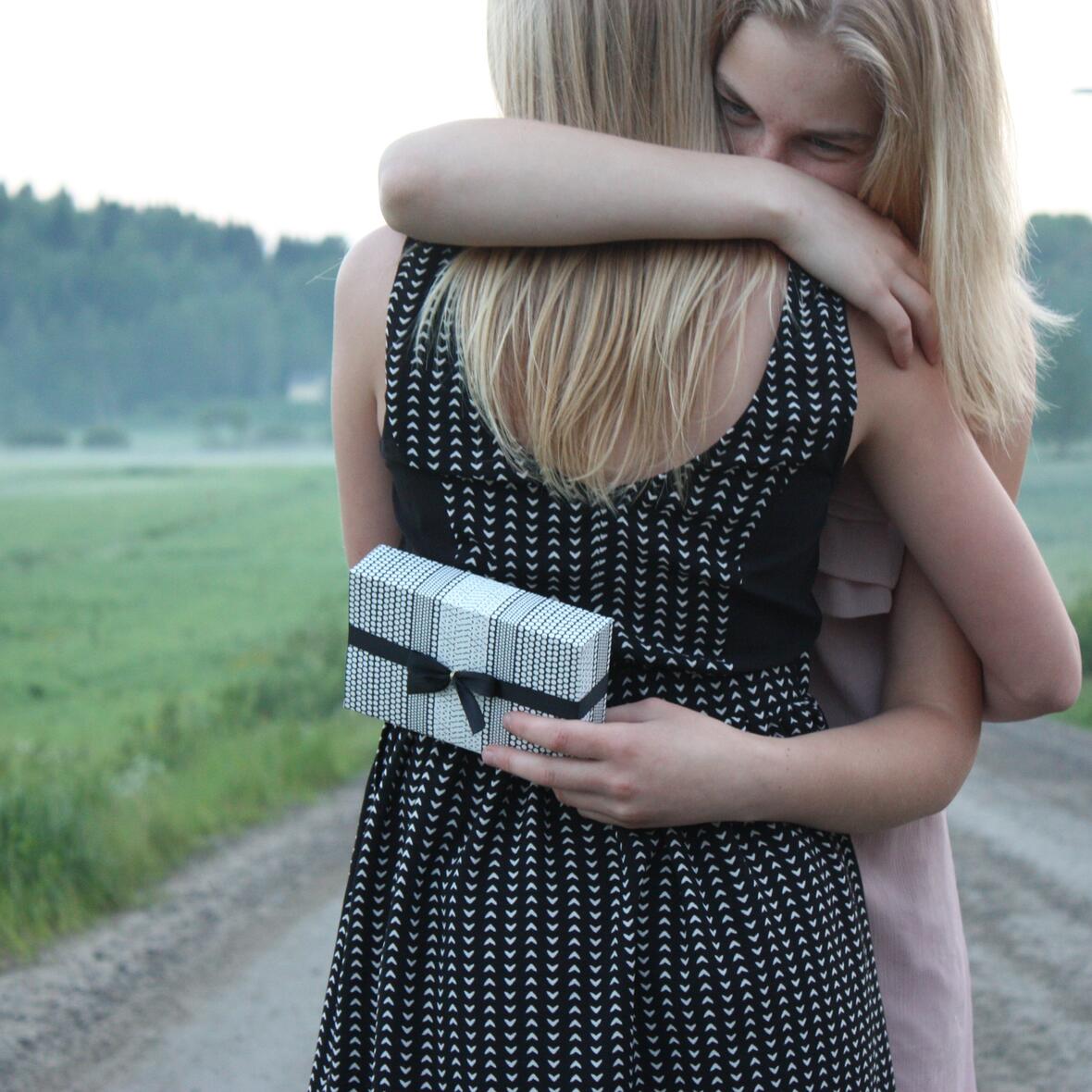 Zwei Frauen umarmen sich auf einem Feldweg. Hinter dem Rücken hält eine ein Geschenk in der Hand. 