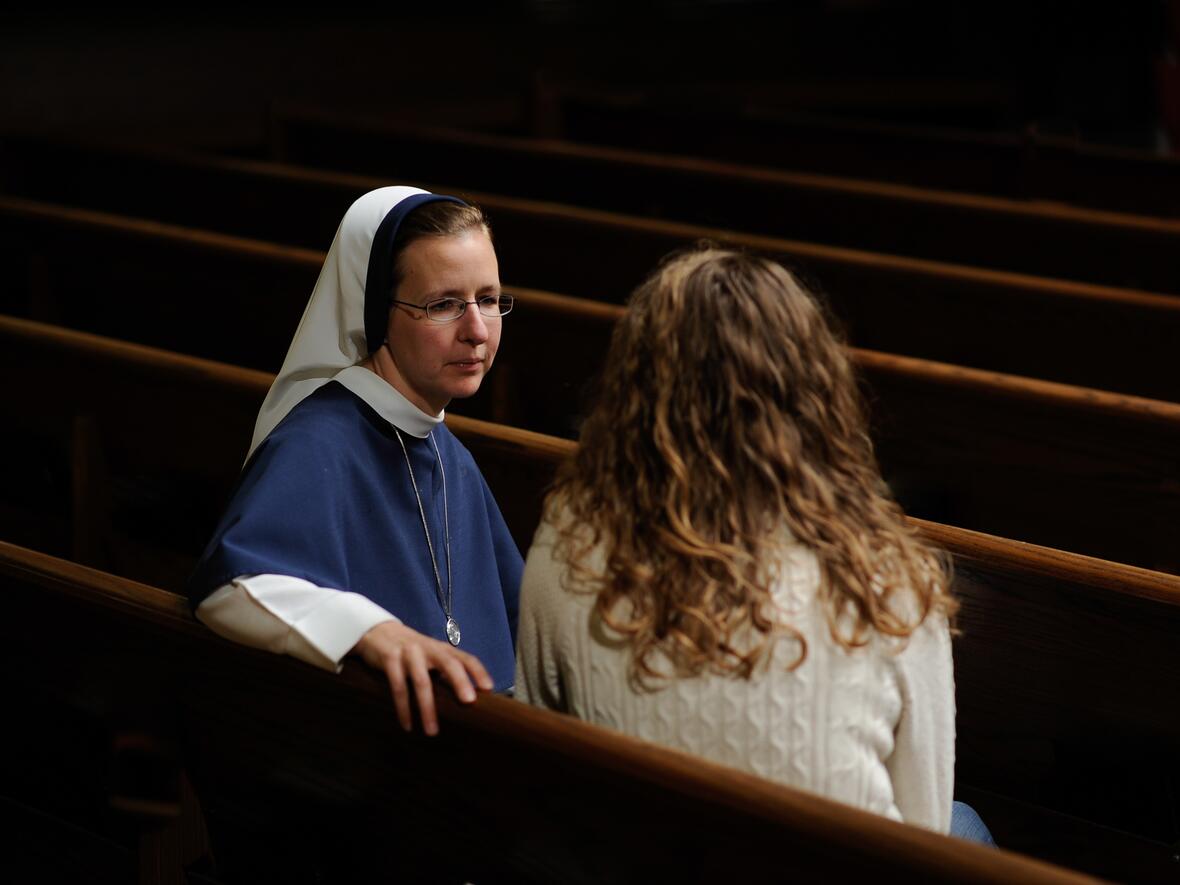 Eine Ordensfrau und eine Frau sitzen in einer Kirchenbank und unterhalten sich. 