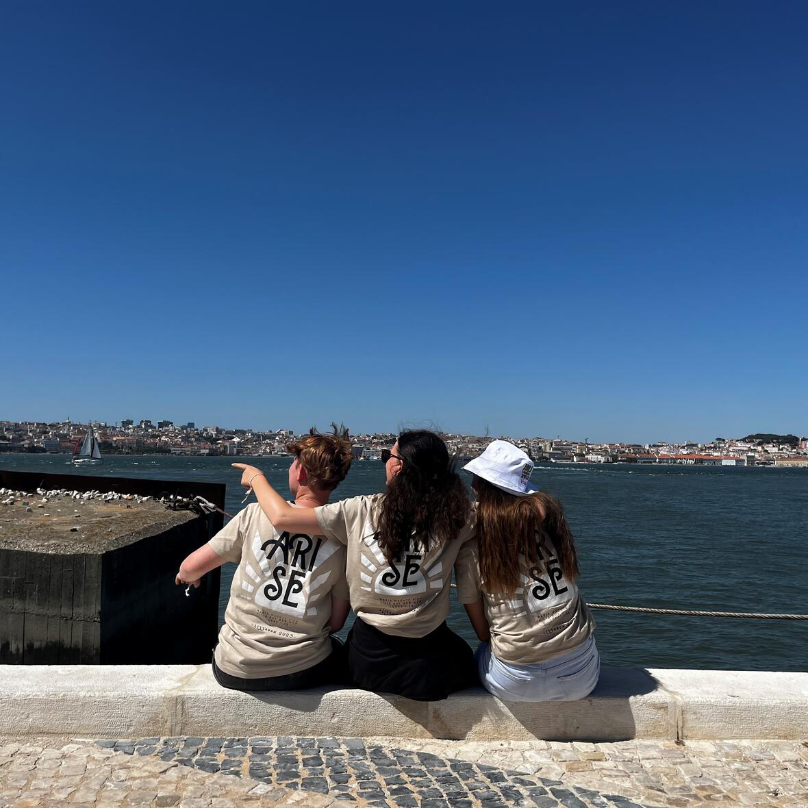 Drei junge Frauen mit hellbraunen T-Shirts sitzen am Ufer eines Gewässers