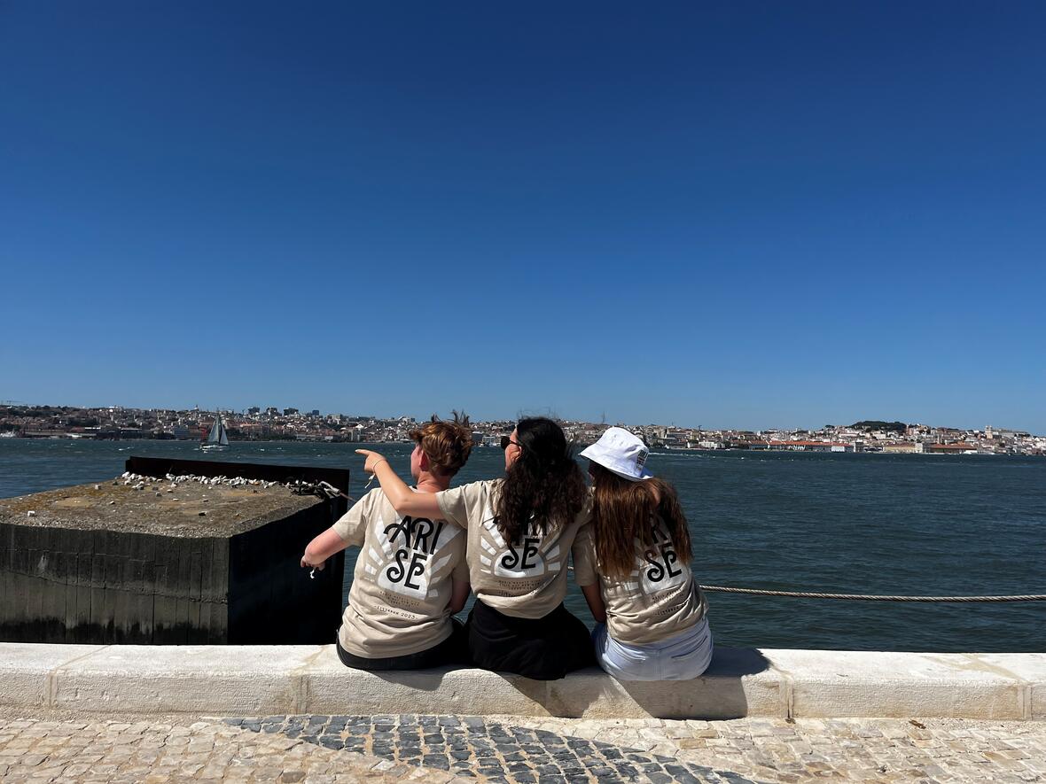 Drei junge Frauen mit hellbraunen T-Shirts sitzen am Ufer eines Gewässers