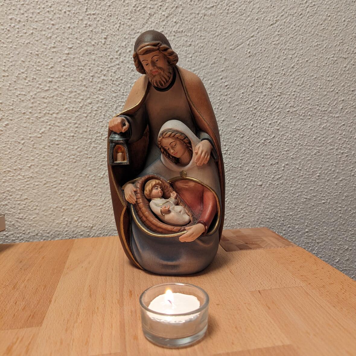 Blockfigur aus Holz mit einem Mann der eine Laterne hält und seinen Mantel um eine Frau geschlagen hat, die auf ein Kind herab blickt. Davor ein Teelicht. 