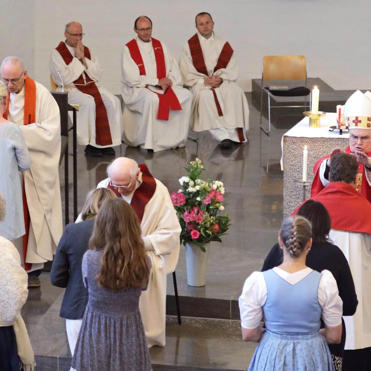 Vorne reihen sich Personen, die von Priestern gesegnet werden, dahinter drei Priester sitzend.