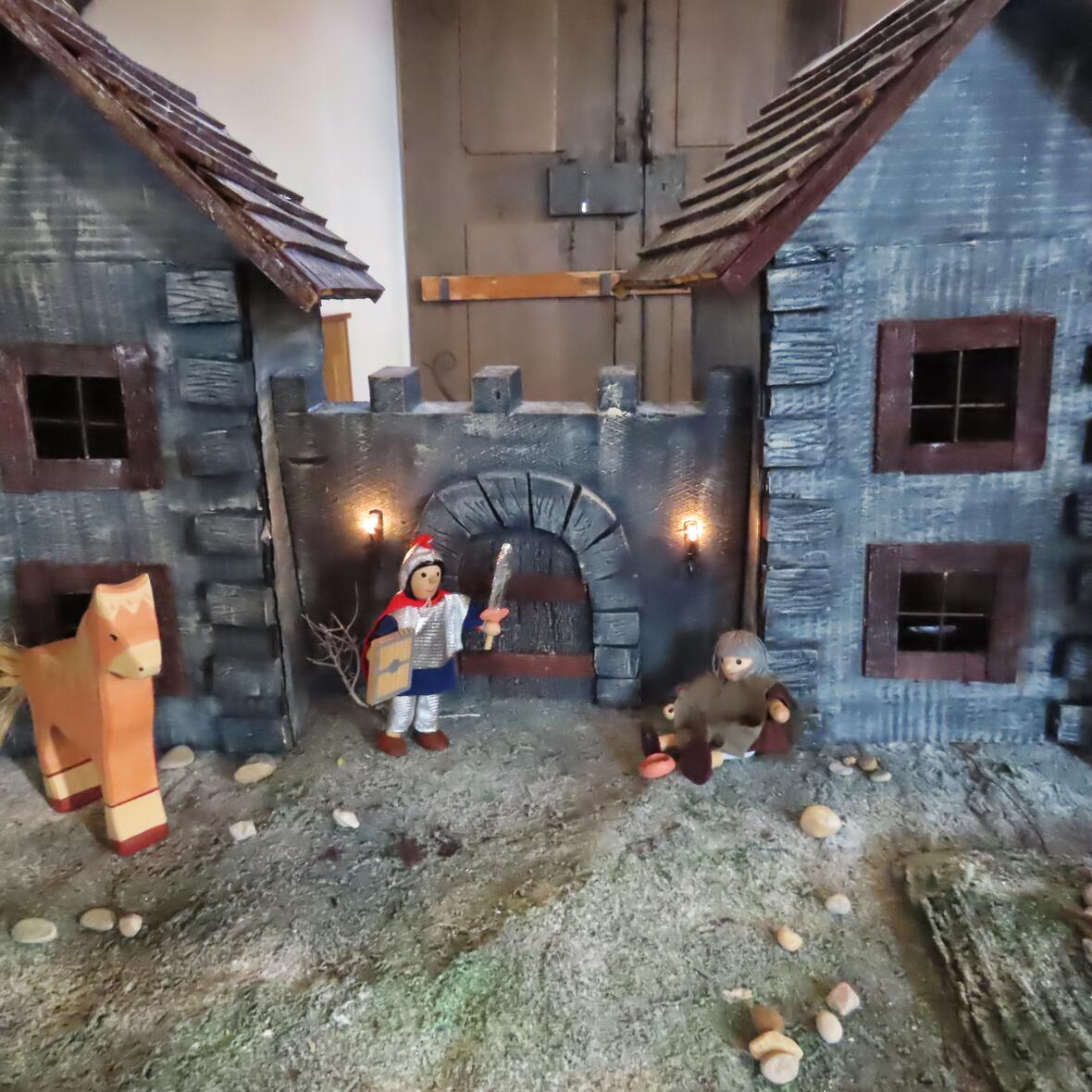 Zwei kleine Häuser mit Schindeldächern und Figuren aus Holz davor, darunter ein Pferd, ein Mensch mit Laterne und eine sitzende Figur