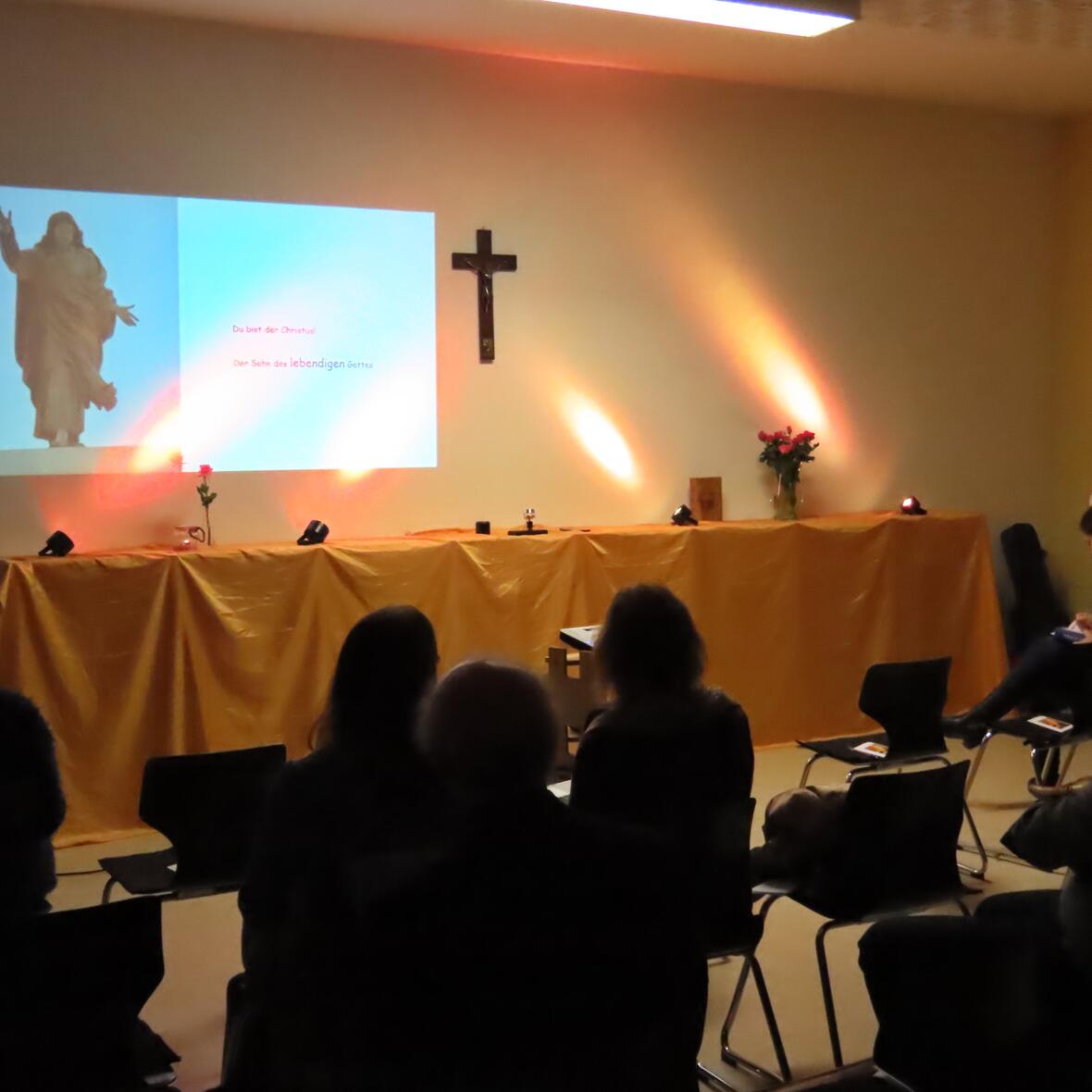 Vortragender steht vor einer Wand mit projiziertem Bild eines Jesus-Statue und Text, an der Wand hängt ein Kreuz, Publikum sitzt auf Stühlen in einem Raum mit gelblich beleuchteter Wand.