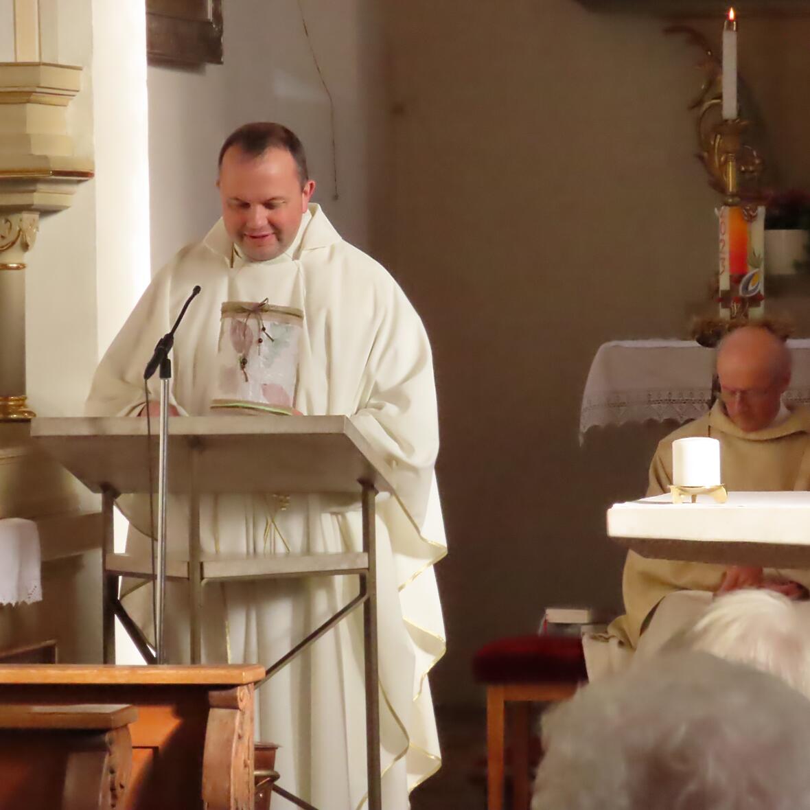 Ein Priester in weißem Gewand steht am Ambo in einer Kirche und spricht. 