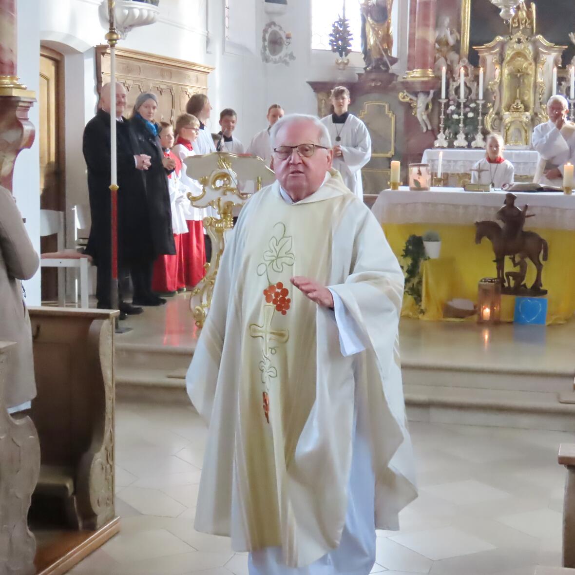 Ein Priester im weißen Gewand spricht gehend im Mittelgang. Im Altarraum hinter ihm sind Menschen unterschiedlichen Alters zu sehen. 