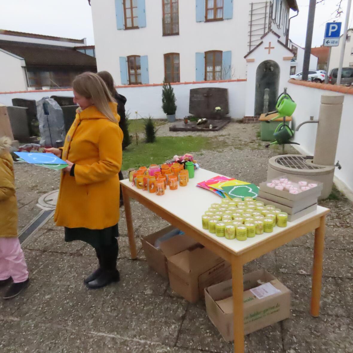 Ein Tisch mit Kerzen steht im Freien. Eine Frau und ein Kind stehen daneben. Eine niedrige Mauer umgibt den Bereich. 