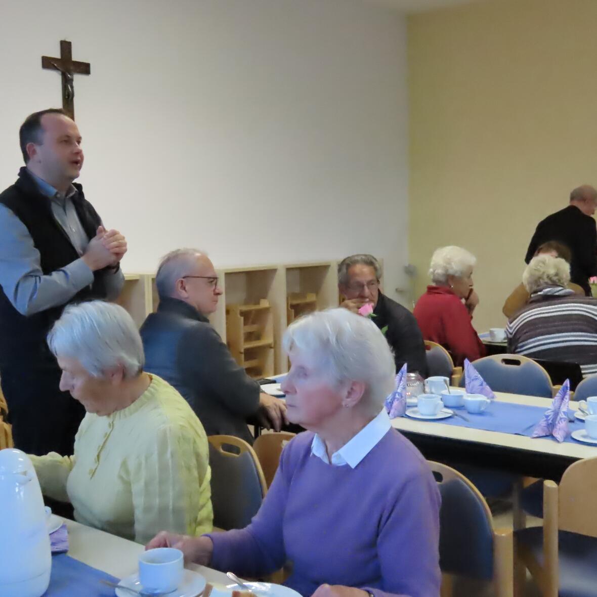 Ein Mann spricht vor einer Gruppe von Senioren. Diese sitzen an gedeckten Tischen bei Kaffee und Kuchen. 