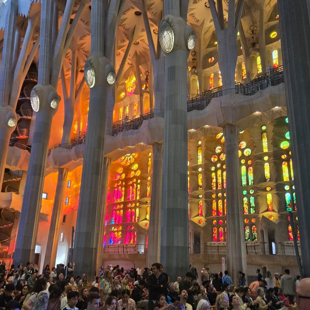 Die linke Seite des Hauptschiffes der Sagrada Familie, das mit vielen Besuchern gefüllt ist: hinter einer Säulenreihe dringt durch die Kirchenfenster Licht in verschiedenen Gelb- und Rottönen.