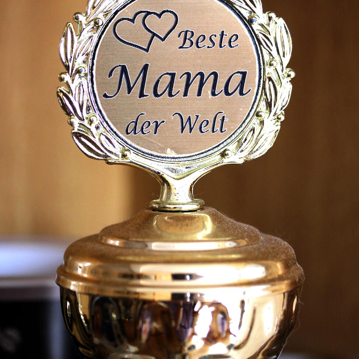silberfarbener Pokal, auf dessen Deckel ein Schild mit der Aufschrift „Beste Mama der Welt“ und zwei kleinen Herzen links oben befestigt ist. 