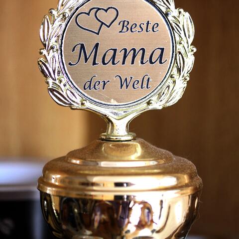 silberfarbener Pokal, auf dessen Deckel ein Schild mit der Aufschrift „Beste Mama der Welt“ und zwei kleinen Herzen links oben befestigt ist. 
