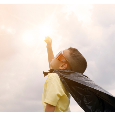 Kleiner Junge mit gelbem Hemd, dunklem Helden-Umhang und übergroßer Sonnenbrille streckt die rechte Hand zum Himmel, der Sonne entgegen.  