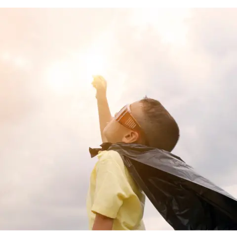 Kleiner Junge mit gelbem Hemd, dunklem Helden-Umhang und übergroßer Sonnenbrille streckt die rechte Hand zum Himmel, der Sonne entgegen.  