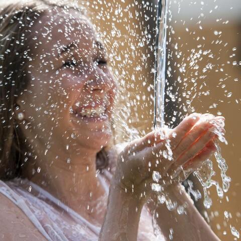 Lächelnde Frau fängt Wasserstrahl mit den Händen auf, Sonnenlicht im Hintergrund.
