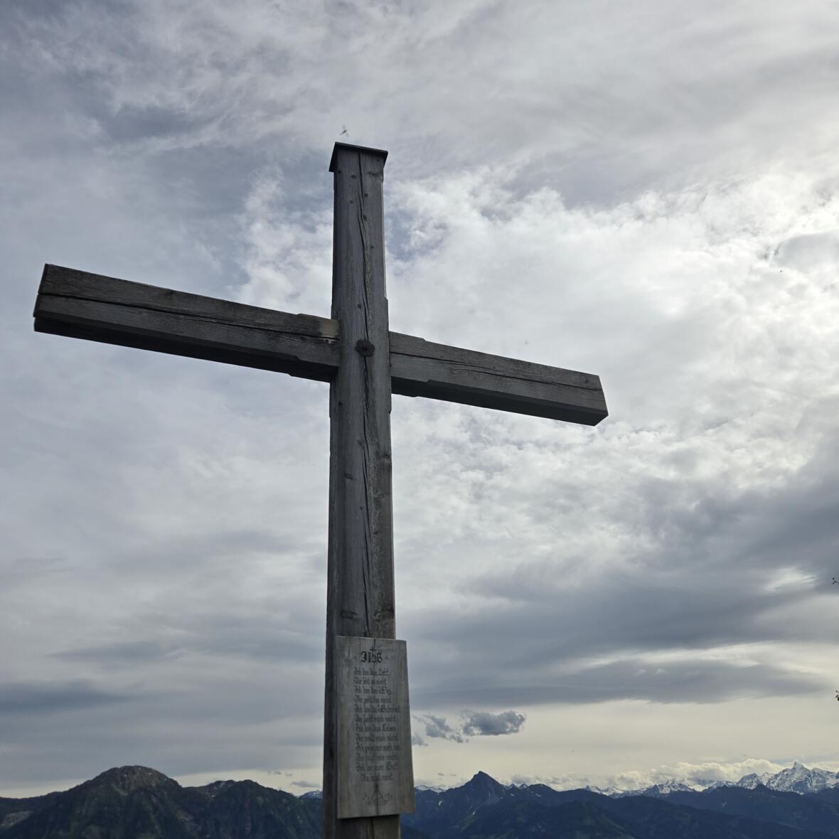 Hölzernes Gipfelkreuz des Hochglockers in Österreich mit einer Holztafel, auf der ein Gebet steht, im Hintergrund blauer wolkenverhangener Himmel über einer Bergkette