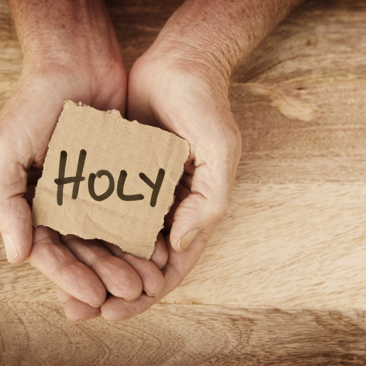 Zwei Hände liegen als Schale geformt auf einer Holzplatte. In den Handflächen liegt ein Stück Karton mit dem englischen Wort Holy