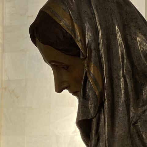 Eine Skulptur von Maria. Ihr Gesicht liegt im Schatten. Der Hintergrund ist erhellt. 