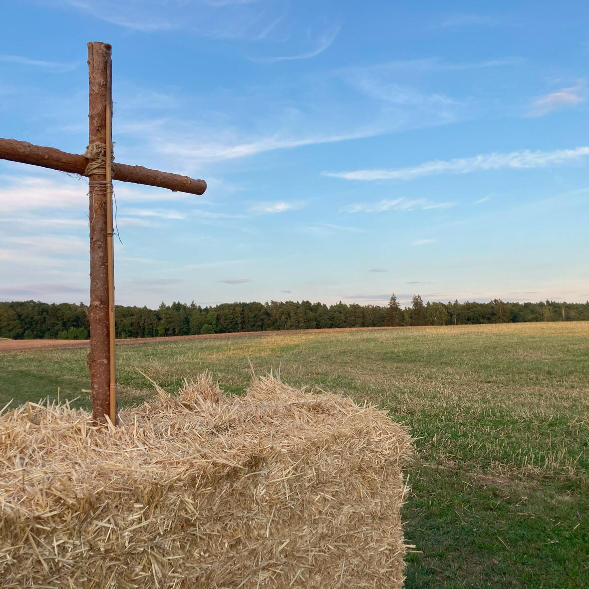 Gott im Museum Auf dem Bild ist eine abgeerntete Landschaft dargestellt. Auf einem Strohballen ist ein Kreuz zu sehen.