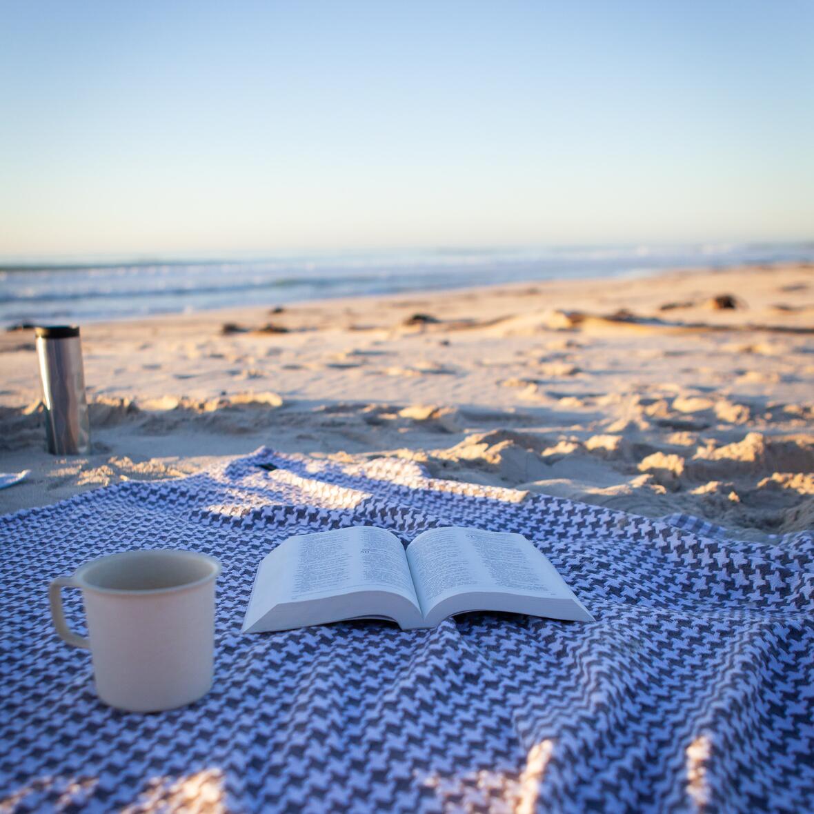 Ein offenes Buch und eine Tasse auf einer Decke am Strand.