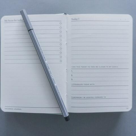 Auf einem grauen Untergrund liegt ein aufgeschlagenes Notizbuch auf dem ein grauer Stabilostift liegt.