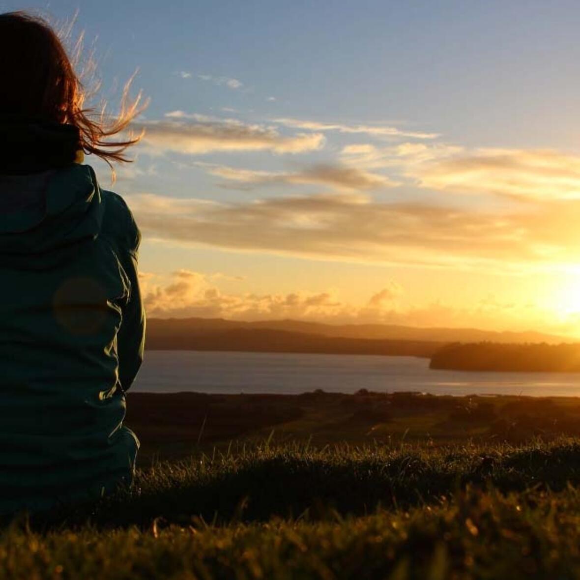 Frau von hinten sitzt auf einer Wiese bei einem Sonnenuntergang