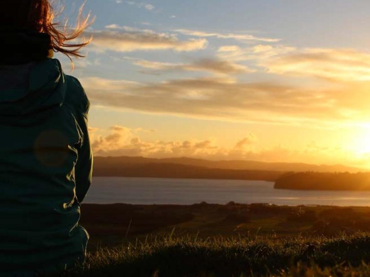 Frau von hinten sitzt auf einer Wiese bei einem Sonnenuntergang