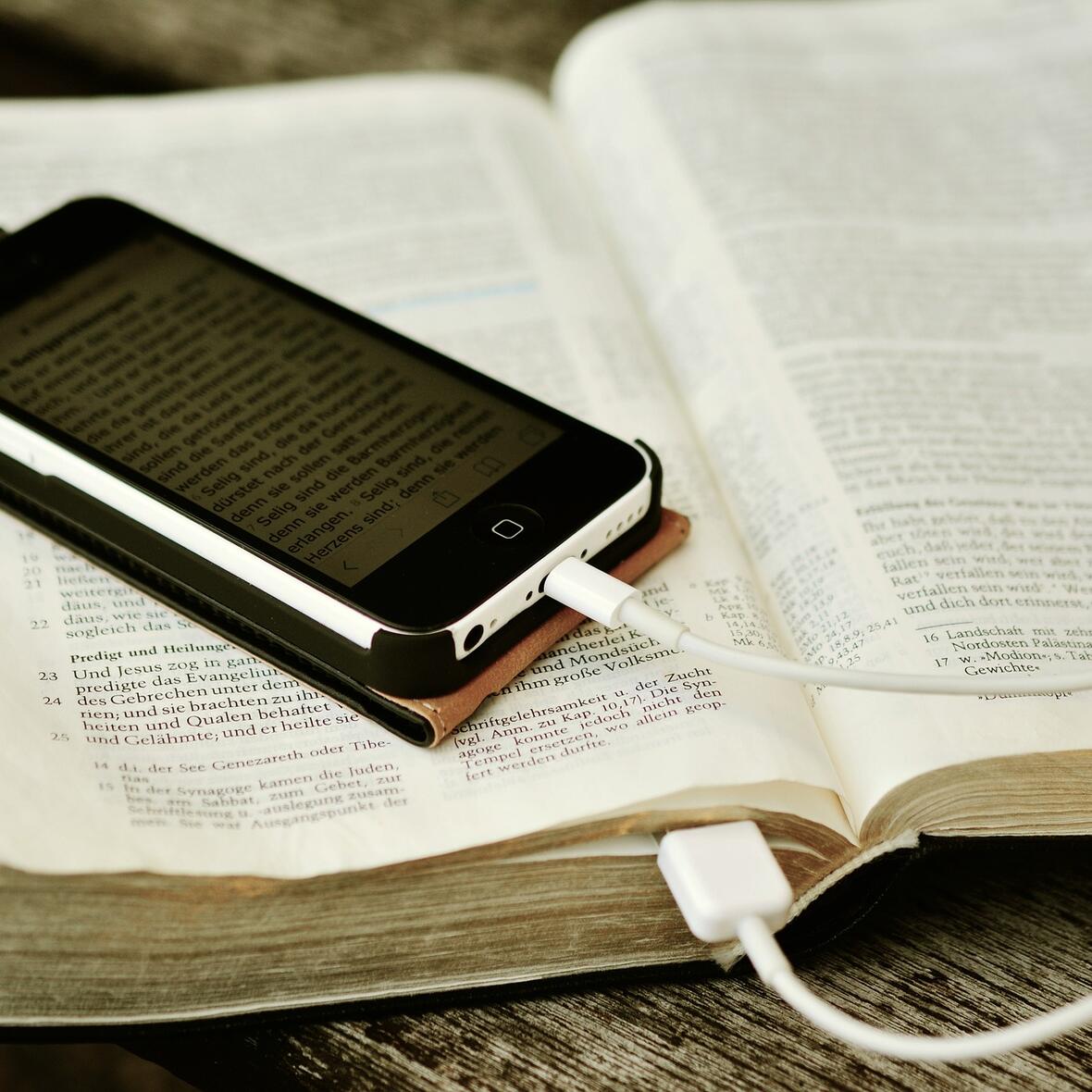 Auf einer Bibel, die auf einem Tisch liegt, befindet sich ein Handy. Das Ladekabel steckt in der Bibel 
