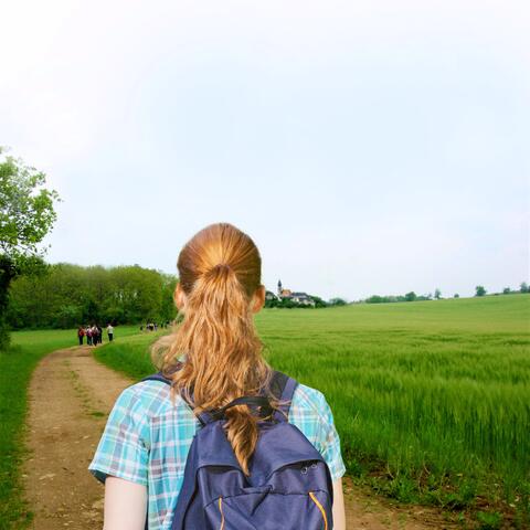 Eine junge Frau geht auf einem Feldweg an grünen Feldern entlang auf ein Dorf zu. 