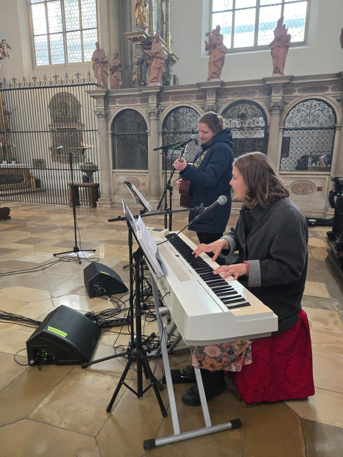 Eine junge Frau sitzt am E-Piano während sie von einer jungen Erwachsenen mit der Gitarre begleitet wird. 