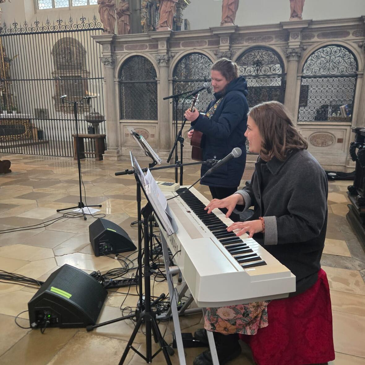Eine junge Frau sitzt am E-Piano während sie von einer jungen Erwachsenen mit der Gitarre begleitet wird. 