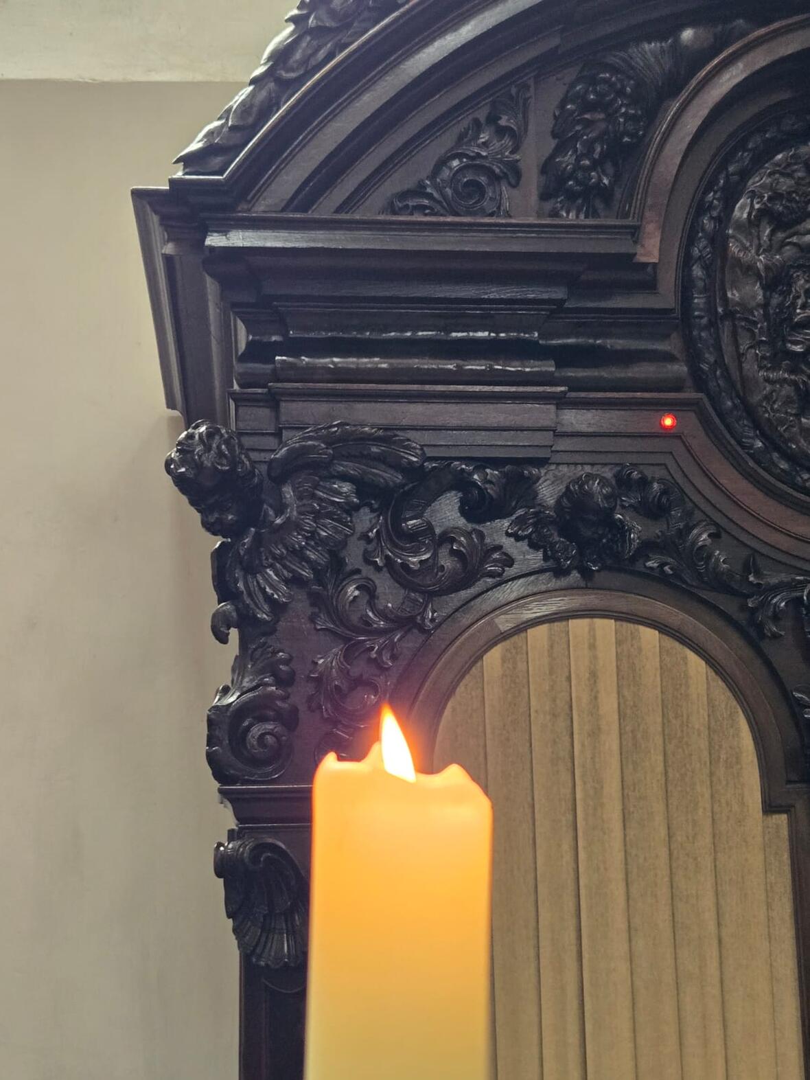 Eine brennende Kerze vor einem Beichtstuhl in dem gerade gebeichtet wird. 