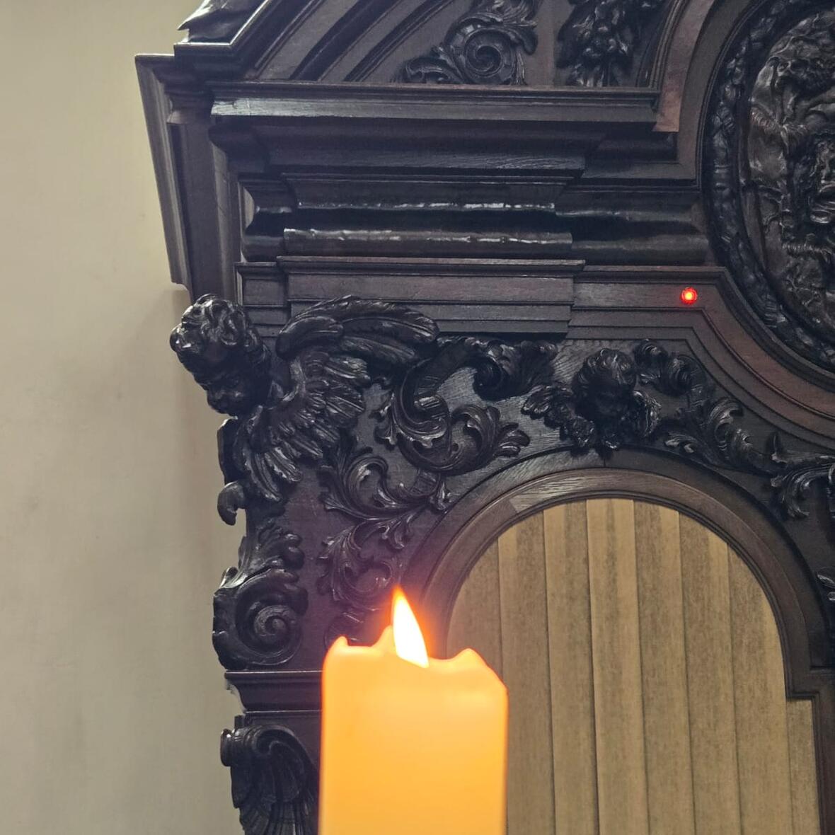 Eine brennende Kerze vor einem Beichtstuhl in dem gerade gebeichtet wird. 