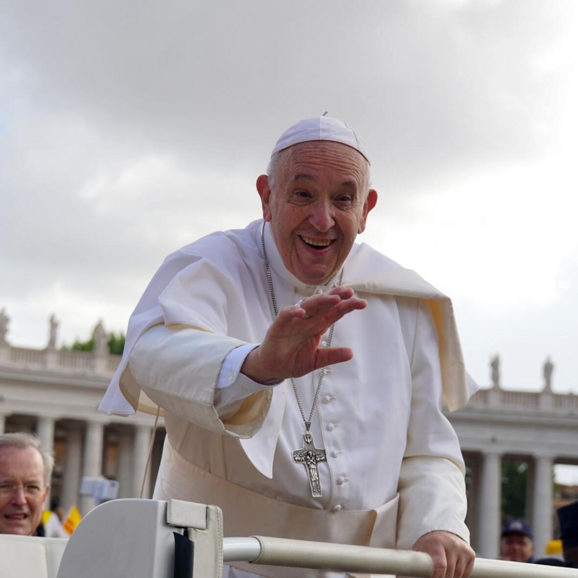 Papst Franziskus wendet sich bei der Generalaudienz der Menschenmenge lächelnd zu und streckt dem Betrachter die Hand entgegen. 
