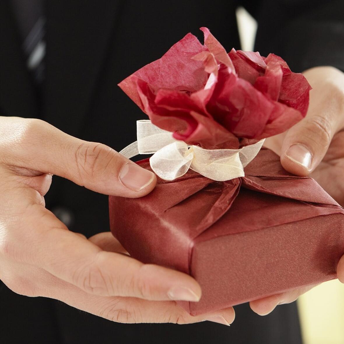 Eine dunkel gekleidete Person übergibt mit beiden Händen ein rotes Geschenk mit Schleife. 