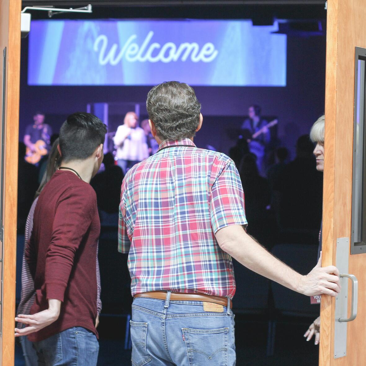 Zwei Männer betreten durch eine Flügeltür einen Saal. Im Hintergrund steht das englische Wort: Welcome!