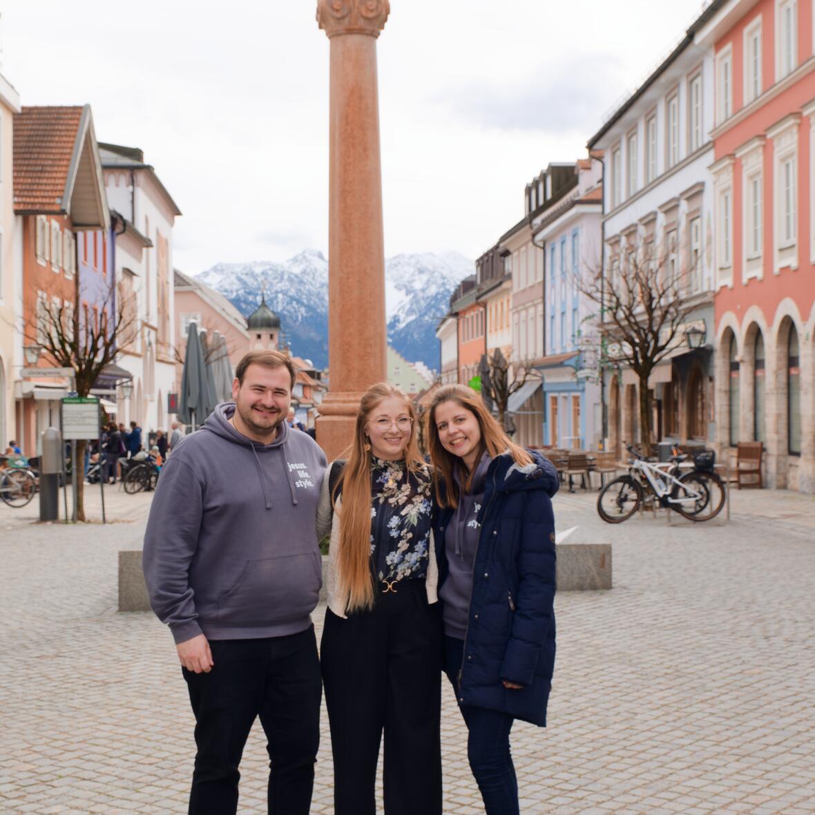 Junge Erwachsene stehen lächelnd vor der Säule auf dem Marktplatz in Murnau. 