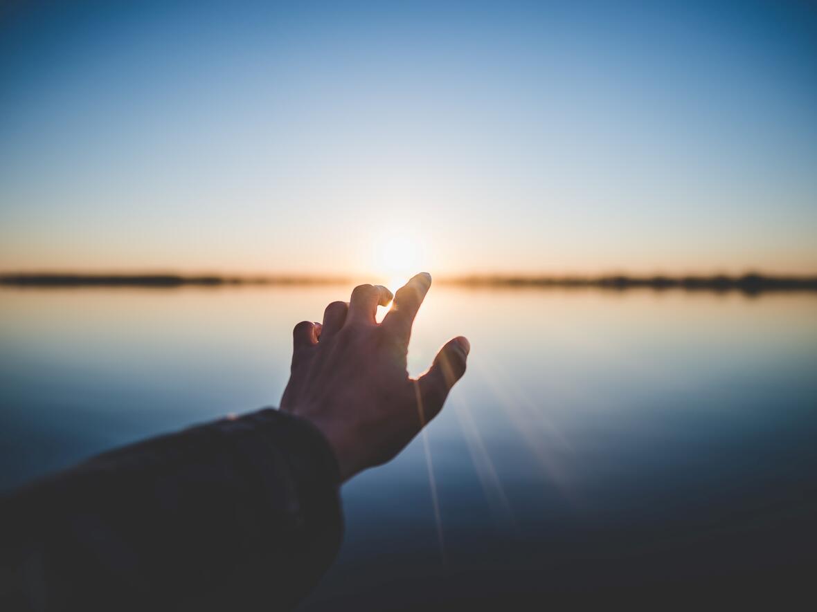 Eine Hand, die auf den Horizont zeigt. Im Hintergrund geht die Sonne auf