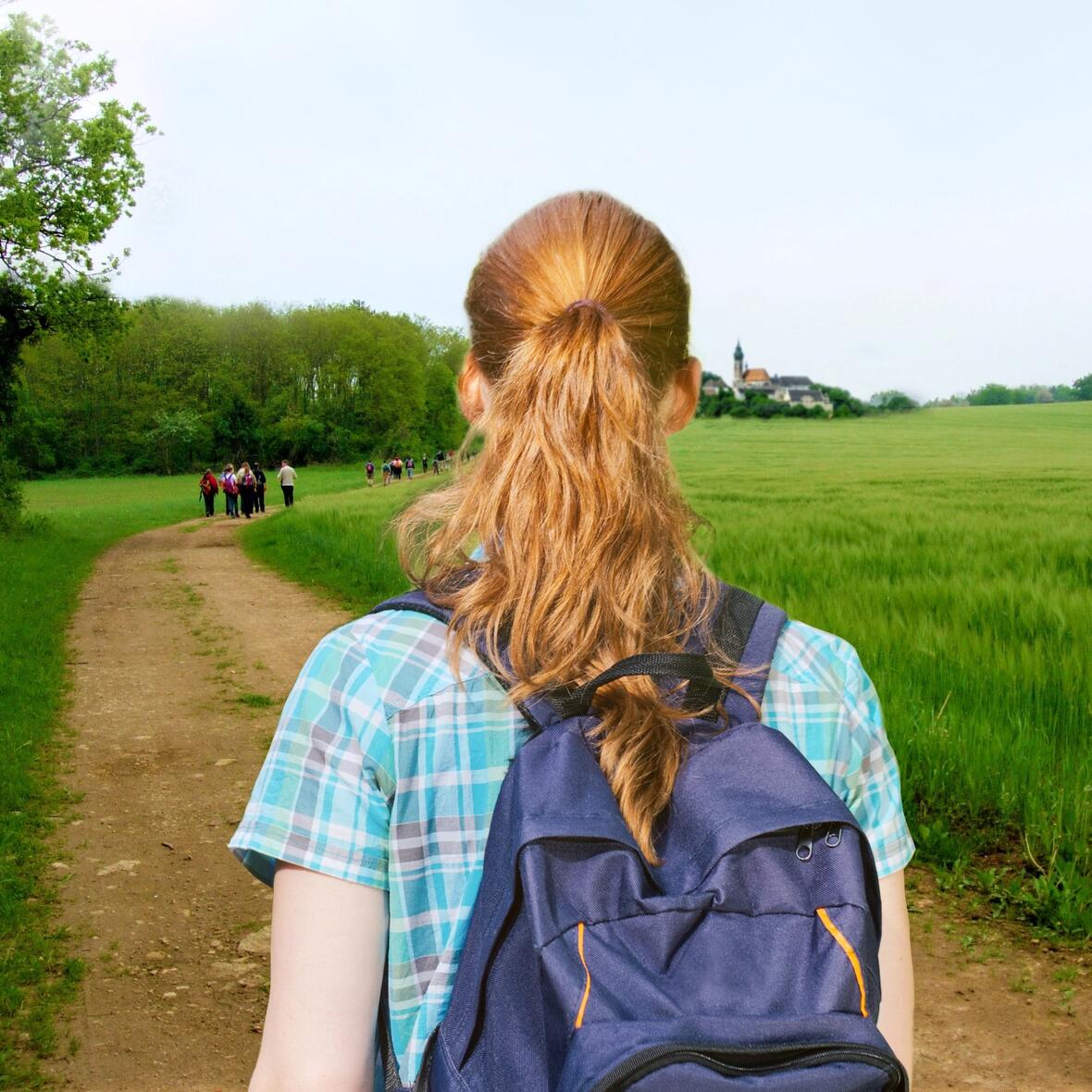 Eine junge Frau geht auf einem Feldweg an grünen Feldern entlang auf ein Dorf zu. 