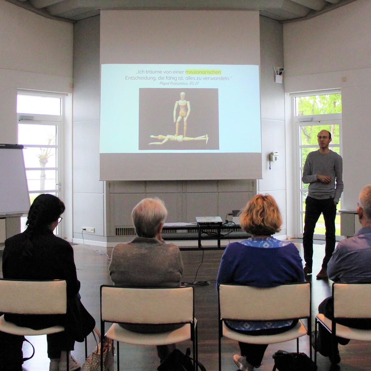 Der Referent spricht vor einer Gruppe Menschen. Im Hintergrund ist eine Powerpoint zu sehen